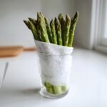 Fresh asparagus spears standing upright in a glass of water, wrapped in a damp paper towel, illustrating how to keep produce fresh.