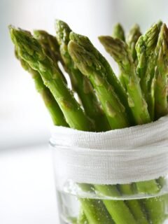 Fresh green asparagus spears standing upright in a glass of water, wrapped with a white cloth, demonstrating how to keep produce fresh.
