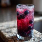 Close-up of a tall glass filled with a vibrant red Blueberry Blackberry Mocktail, ice, and whole berries.