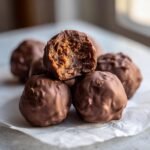 A stack of rich, chocolate-coated Butterfinger Balls, with one candy on top bitten open to show the textured, peanut-buttery center.