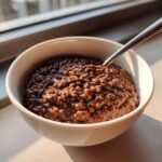 A close-up of creamy, dark brown Chocolate Rice Pudding served in a white bowl with a spoon resting inside.