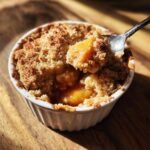 A spoonful of warm Cinnamon Peach Crumble being lifted from a white ramekin onto a wooden surface.