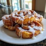Close-up of several golden Cinnamon Roll French Toast Bites drizzled heavily with white icing on a white plate.