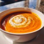 Close-up of a bowl of vibrant orange soup, swirled with cream and topped with black pepper, perfect for warm cozy fall recipes.