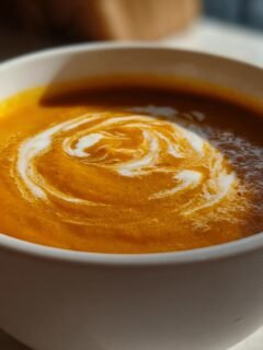 Close-up of a bowl of vibrant orange, creamy soup garnished with a swirl of white cream, perfect for warm cozy fall recipes.