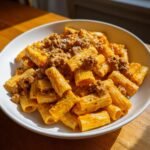 A close-up of a white bowl filled with rich, orange-hued Creamy Sausage Rigatoni, topped with crumbled sausage.