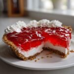 A slice removed from a vibrant red Easter Jell O Pie featuring a creamy white layer and graham cracker crust, topped with whipped cream.