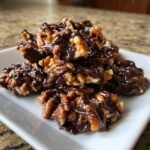 A stack of Easy Chocolate Caramel Pecan Clusters drizzled generously with melted dark chocolate on a white serving dish.