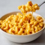 A spoonful of creamy, bright orange Easy Vegan Pumpkin Mac Cheese being lifted from a white bowl.