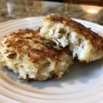 Two golden-brown, pan-fried crab cakes on a white plate, one broken open to show the flaky white crab meat inside.