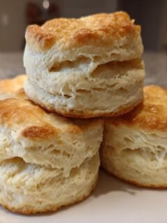 Close-up of three golden-topped, fluffy Homemade Buttermilk Biscuits stacked on a white plate.