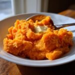 A close-up of fluffy, bright orange Mashed Sweet Potatoes served in a white bowl with a pat of melting butter on top.