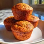 A close-up stack of four golden brown Sweet Potato Muffins resting on a white plate, highlighted by sunlight.