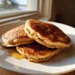 A stack of three golden-brown Healthy Vegan Flourless Apple Pancakes drizzled with syrup on a white plate.