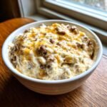 A close-up of a creamy Hot Ranch Sausage Dip topped with melted cheddar cheese in a white bowl.