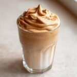 A tall glass filled with milk and topped with a dramatic swirl of whipped Iced Dalgona Coffee Vanilla Latte foam.
