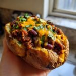A person holding a baked potato filled with spicy bean chili, melted cheddar cheese, and fresh cilantro, representing Loaded Potatoes With Spicy Beans.