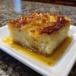 A square serving of moist Magic Lemon Cobbler with a caramelized top, sitting in a pool of lemon sauce.