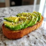 Close-up of Mind Blowing Avocado Toast featuring perfectly fanned avocado slices seasoned with flaky salt and pepper on toasted bread.