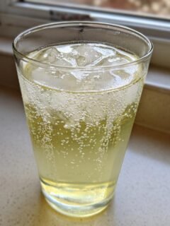 A tall glass filled with bubbly Natural Homemade Lemon Ginger Ale and ice cubes, sitting on a light countertop near a window.