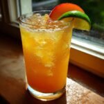 A tall, cold glass filled with a vibrant orange Peachy Ginger Beer Mocktail, garnished with peach and lime slices.