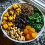 Overhead view of a colorful bowl featuring iron-rich plant based foods like tofu, spinach, quinoa, cashews, and dried fruit.