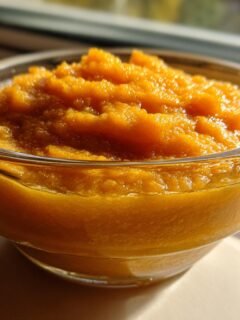 A clear glass bowl filled with vibrant orange homemade pumpkin puree, sitting in bright sunlight.