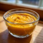 A small glass bowl filled with smooth, bright orange pumpkin puree, sitting on a wooden surface.