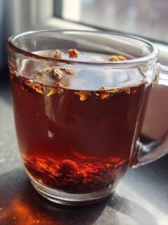 A clear glass mug filled with dark, rich Roasted Dandelion Strawberry Tea, showing steeped ingredients at the bottom.