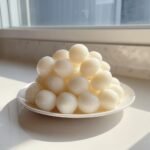 A mound of glossy, white spherical yogurt recipe balls piled high on a small white plate near a window.