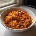 A white bowl filled with saucy, curly noodles, shredded chicken, and sliced carrots, representing Spicy Chili Garlic Ramen With Chicken Veggies.