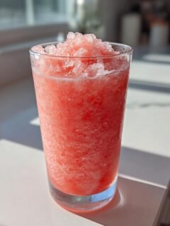 A tall glass filled with a bright pink, icy Watermelon Slushie topped with crushed ice.