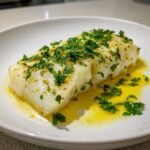A close-up of a tender Baked Lemon Butter Cod fillet topped with fresh parsley and resting in lemon butter sauce.