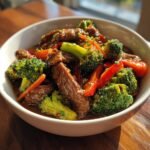 A close-up of a serving of Beef Stir Fry With Vegetables featuring tender beef strips, bright green broccoli, and red bell peppers in a rich brown sauce.