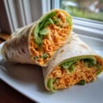 A Buffalo Chicken Salad Wrap cut in half diagonally, showing creamy orange filling and crisp lettuce inside a tortilla.