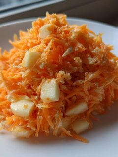 A close-up of a serving of bright orange Carrot Apple Salad with visible chunks of pale apple on a white plate.