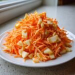 A mound of bright orange shredded carrots mixed with diced white apple pieces, forming a fresh Carrot Apple Salad on a white plate.
