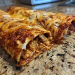 Two freshly baked Bean And Cheese Enchiladas covered in melted red sauce and shredded cheese on a granite countertop.