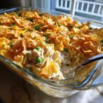 A serving spoon scoops into a glass dish of creamy Tuna Noodle Casserole topped with crushed chips and peas.