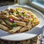 A close-up of creamy Tuna Pasta Recipe served on a white plate, topped with chopped red onion and green onions.