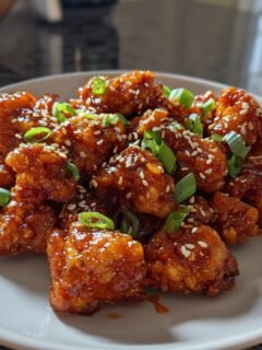 A plate piled high with glossy, saucy Firecracker Chicken pieces, garnished with sesame seeds and chopped green onions.