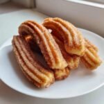 A stack of freshly fried, ridged churros coated generously in cinnamon sugar on a white plate.
