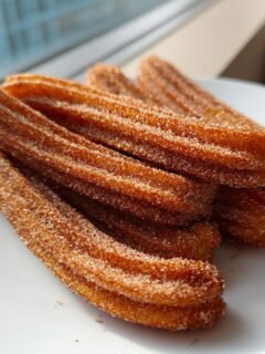 A pile of freshly fried, cinnamon-sugar coated churros resting on a white plate indoors.