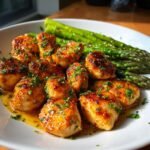 Close-up of golden brown Garlic Butter Chicken Bites drizzled with sauce and parsley, served alongside bright green asparagus.