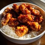 Close-up of crispy chicken pieces coated in sticky honey garlic sauce served over white rice in a bowl.