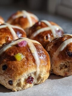 A close-up of several freshly baked, glossy Hot Cross Buns With Candied Fruit, featuring white icing crosses and colorful fruit pieces.