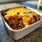 A square white baking dish filled with baked Mexican Quinoa Casserole topped with melted cheddar cheese.