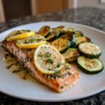 A cooked fillet of One Pan Lemon Herb Salmon And Zucchini topped with lemon slices and herbs, served with roasted zucchini rounds.