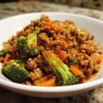Close-up of Orange Ground Chicken stir-fry mixed with bright green broccoli florets and diced carrots in a white bowl.