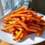 A close-up of a white plate piled high with crispy, golden Oven Baked Sweet Potato Fries.